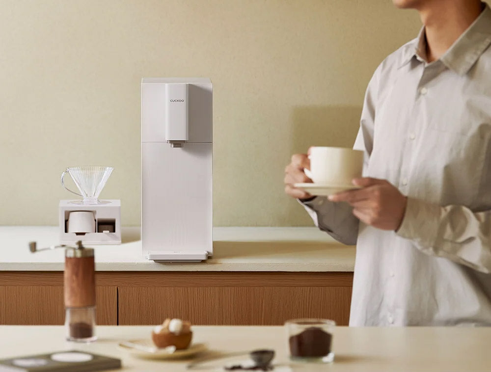 A person in a light gray shirt holds a white cup on a counter beside a STEAM 100 Barista water purifier and a small pour-over dripper, with coffee beans and a grinder in the foreground.