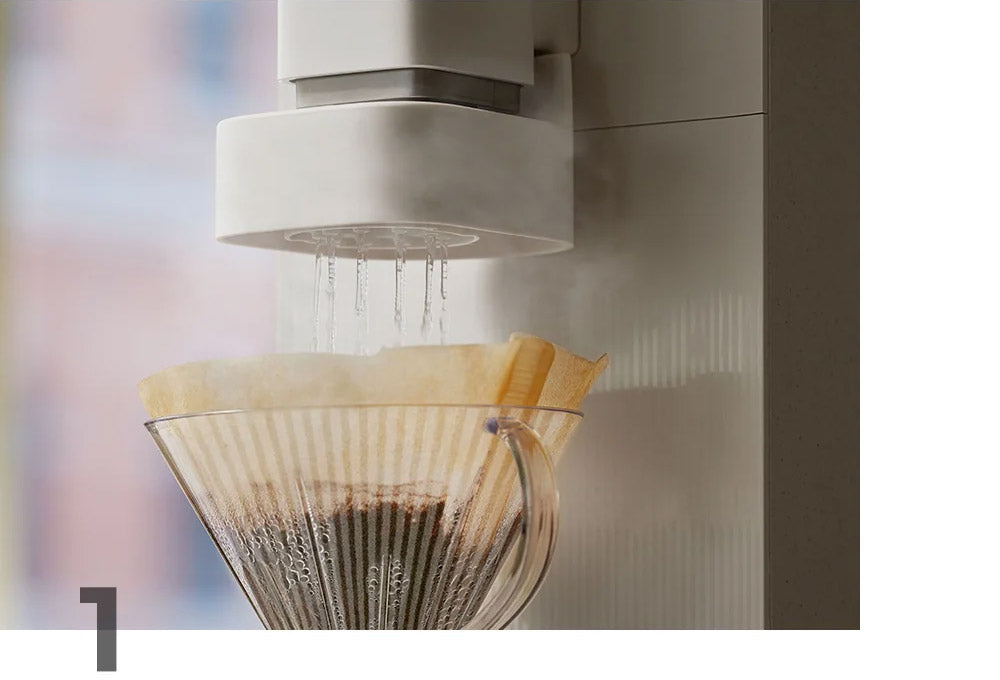 A close-up of the STEAM 100 Barista's drip module with water dripping from a the module into a clear glass cone filter containing coffee grounds and a beige paper filter.