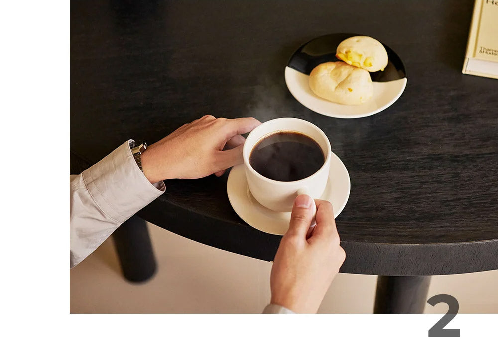 A person's hands holding a white ceramic cup of black coffee on a matching saucer, set on a dark wooden table next to a plate of pastries.