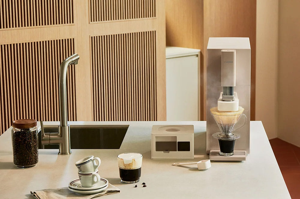 A STEAM 100 Barista water purifier setup on a light countertop in a minimalist kitchen-like interior. Visible items include a tall white and clear coffee maker with a glass cone filter, a small white box, a glass jar of coffee beans, two stacked ceramic cups, and a small glass cup with dark coffee.
