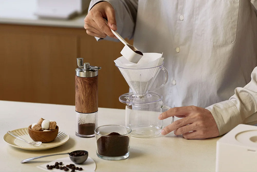 A person in a light beige shirt is shown pouring coffee grounds from a small spoon into a white cone filter on a glass pour-over dripper. The setup is on a light beige countertop with a glass carafe, a wooden-and-silver coffee grinder, a small bowl of sugar and cookies, and coffee beans.