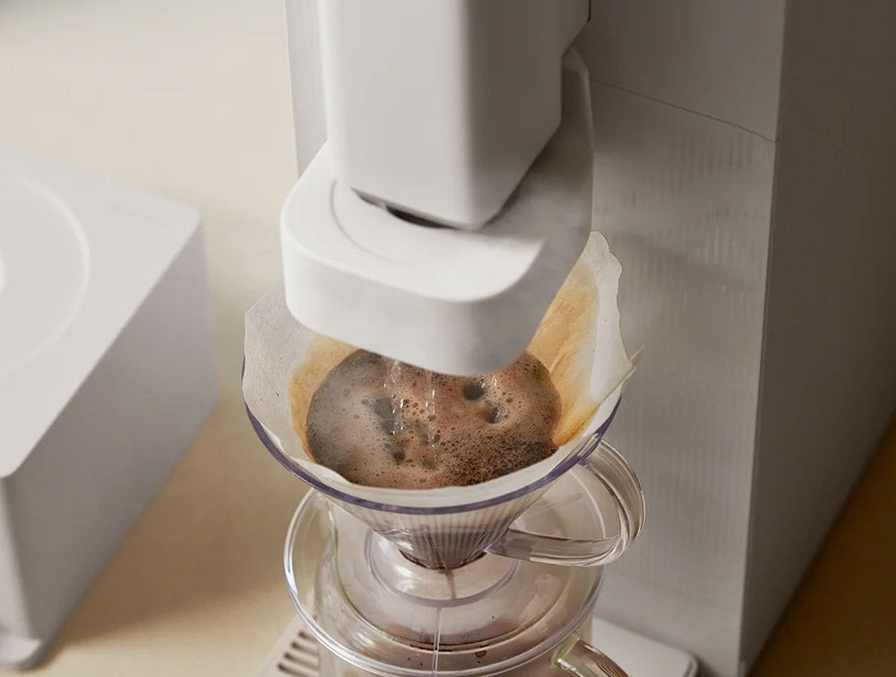 A close-up of STEAM 100 Barista water purifier dripping freshly brewed coffee into a glass carafe, with a white paper filter cone in the cone holder.
